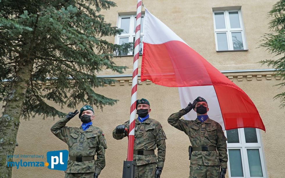 2.05.2021 r. Dzień Flagi Rzeczypospolitej Polskiej; trzech żołnierzy 18. Łomżyńskiego Pułku Logistycznego stoi przy maszcie flagowym, środkowy żołnierz przy pomocy korby wciąga biało-czerwoną flagę na maszt, pozostali - salutują